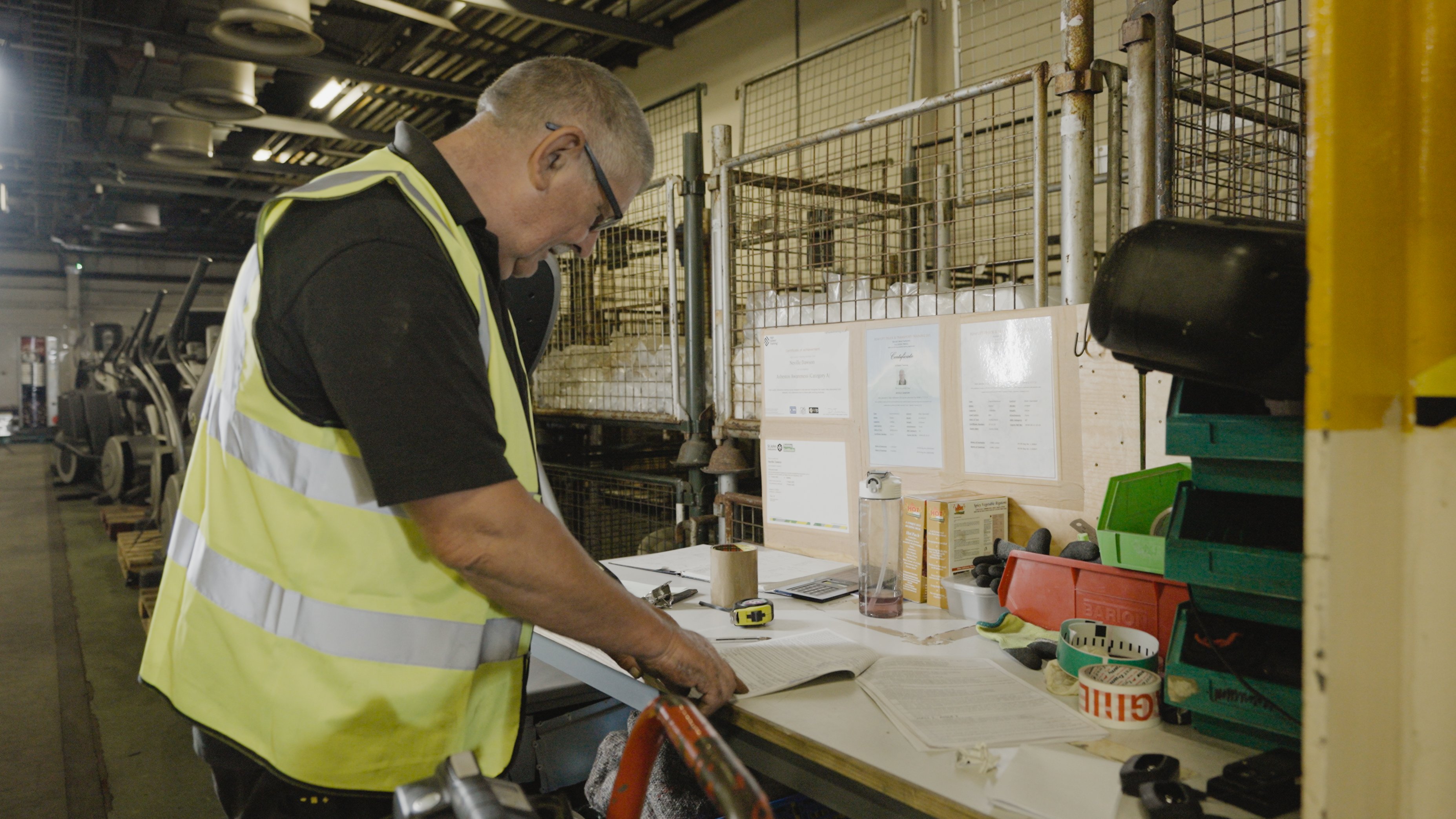 A man wearing a high-viz jacket stood over a work desk in a warehouse, surrounded by surplus assets being prepared for sale.