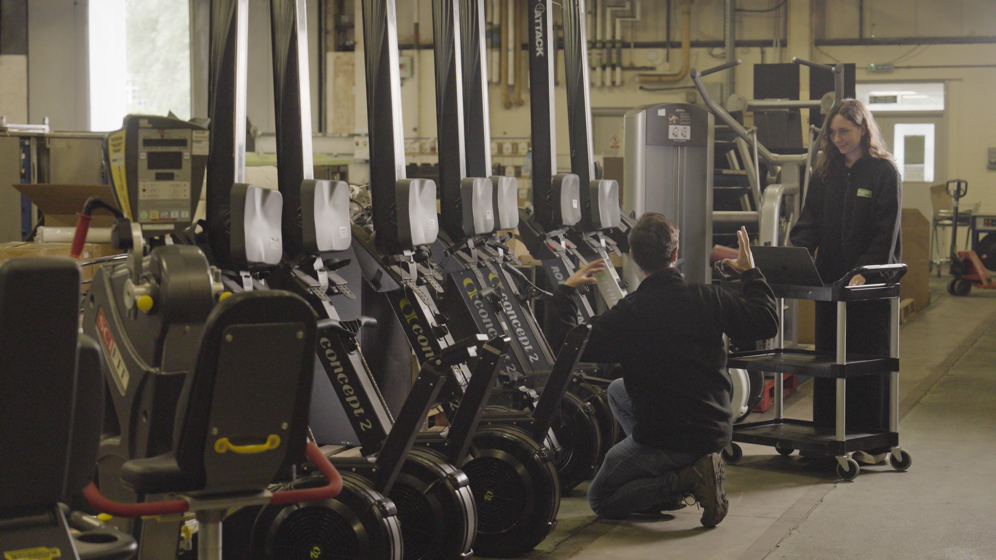A man kneeled on the floor and a woman stood on a computer talking. Surrounded by surplus assets, including gym equipment, being prepared for resale and reuse.